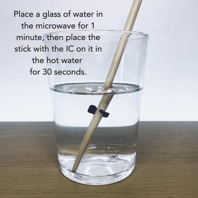 May include: A clear glass of water with a wooden stick and a black clip inside. Text on the image reads: Place a glass of water in the microwave for 1 minute, then place the stick with the IC on it in the hot water for 30 seconds.