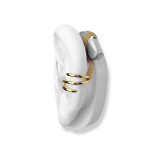 May include: A gold-colored ear cuff with a triple-band design is displayed on a white ear model. The cuff wraps around the ear's cartilage. A silver hearing aid is also visible.