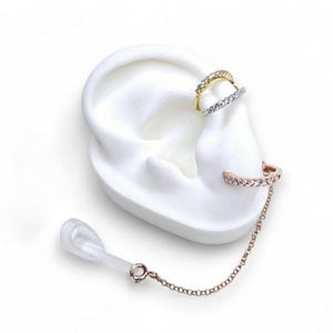 May include: Three ear cuffs on a white ear model. The top cuff is gold-colored with clear stones, the middle cuff is silver-colored with clear stones, and the bottom cuff is rose gold-colored with clear stones and a chain.