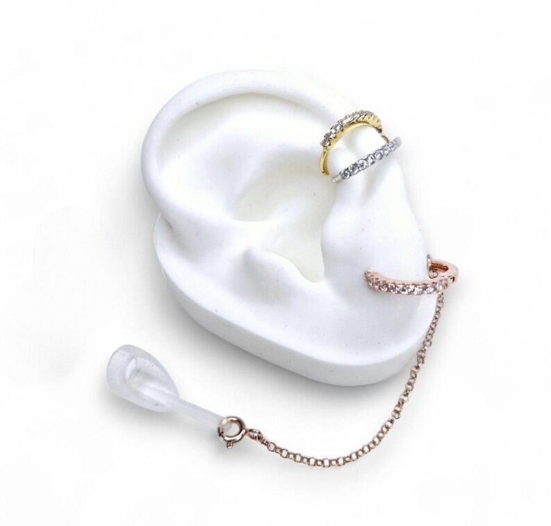 May include: Three ear cuffs on a white ear model. The top cuff is gold-colored with clear stones, the middle cuff is silver-colored with clear stones, and the bottom cuff is rose gold-colored with clear stones and a chain.