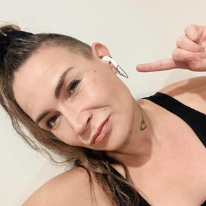 May include: White wireless earbud with a silver clip-on design. The earbud is in the ear of a person. The person is pointing at the earbud with their index finger. The person is wearing a black tank top.
