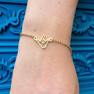 Op de afbeelding: Een goudkleurige armband met een 'shaka' handgebaar bedel. De bedel is verbonden met een fijne ketting. De armband wordt om een pols gedragen, tegen een blauwe achtergrond.