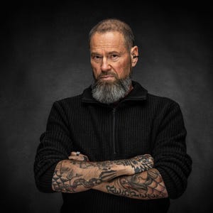 May include: A man with a gray beard and tattoos on his arms, wearing a black zip-up sweater. The background is a dark gray. The man has his arms crossed and is looking directly at the camera.