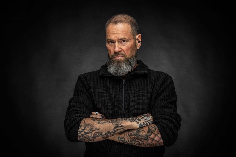 May include: A man with a gray beard and tattoos on his arms, wearing a black zip-up sweater. The background is a dark gray. The man has his arms crossed and is looking directly at the camera.