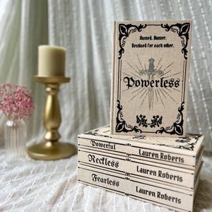 Könnte beinhalten: Ein Stapel von drei Büchern mit den Titeln "Powerful", "Reckless" und "Fearless" von Lauren Roberts. Ein viertes Buch, "Powerless", liegt oben auf. Alle Bücher haben einen beigefarbenen Einband mit schwarzem Text und dekorativen Rändern. Das oberste Buch trägt den Text "Hunted. Hunter. Destined for each other."