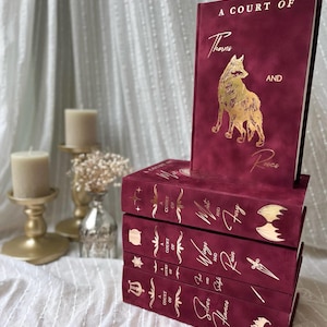 Könnte beinhalten: Ein Stapel weinroter Bücher mit goldenen Buchstaben und Illustrationen. Das oberste Buch zeigt eine goldene Wolfsillustration und den Titel "A Court of Thorns and Roses". Andere Bücher tragen Titel wie "Wings and Ruin" und "Silver Flames".