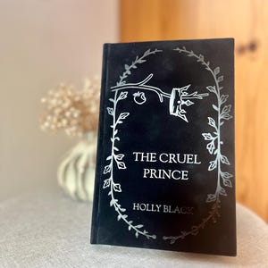 Könnte beinhalten: Ein schwarzes Hardcover-Buch mit dem Titel "The Cruel Prince" von Holly Black. Der Titel und der Name des Autors sind in Silber geschrieben, umgeben von einem floralen Muster. Das Buch steht auf einer neutralen Oberfläche.