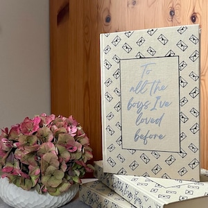 Könnte beinhalten: Bücher mit dem Titel "To All the Boys I've Loved Before" in hellblauer Schrift. Die cremefarbenen Einbände sind mit schwarzen Briefumschlag-Illustrationen versehen. Links steht eine weiße Schale mit rosa und grünen Blumen.
