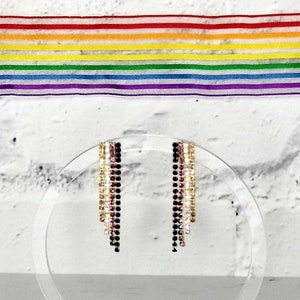 May include: A pair of dangle earrings with black, gold, and pink beads hanging from a clear acrylic stand. The earrings are on a white wooden base.