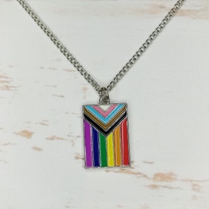 以下が含まれることがあります： カラフルなプライドフラッグのデザインが施された長方形のペンダントが付いたシルバーチェーンネックレス。ペンダントは、上部に茶色、黒、青のシェブロンの形をしたレインボーストライプパターンになっています。