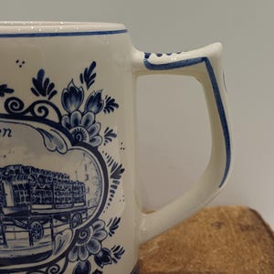 May include: A white ceramic mug featuring blue hand-painted floral motifs and a central oval frame containing a detailed illustration of a horse-drawn carriage. The mug has a sturdy, angular handle with blue decorative trim along the edges.