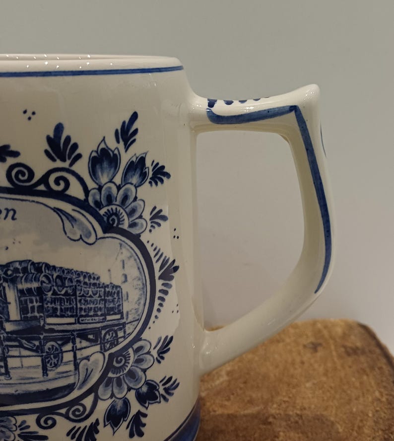 May include: A white ceramic mug featuring blue hand-painted floral motifs and a central oval frame containing a detailed illustration of a horse-drawn carriage. The mug has a sturdy, angular handle with blue decorative trim along the edges.