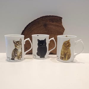 May include: Three white ceramic mugs, each adorned with a different cat illustration. One mug depicts a tabby kitten, another a black cat, and the third a ginger cat. Each mug has a white handle and a signature. The mugs are displayed against a neutral backdrop.