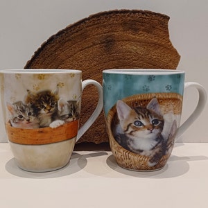 Op de afbeelding: Twee keramische mokken met kattenontwerpen. De ene mok heeft een groep kittens in een mand, terwijl de andere mok een enkel kitten in een mand heeft. Beide mokken hebben een lichtblauwe achtergrond met pootjes.