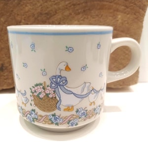 Puede incluir: Una taza de cerámica blanca con un patrón repetitivo de gansos blancos con lazos azules, cestas de mimbre marrón llenas de flores rosas y pequeños detalles florales azules. La taza tiene una línea horizontal azul cerca del borde superior.