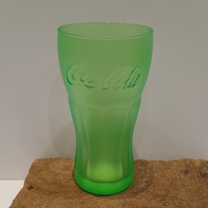 Könnte beinhalten: Ein durchscheinendes, limettengrünes Trinkglas aus Glas mit dem geprägten Coca-Cola-Logo auf der Vorderseite. Das Glas hat eine klassische, geschwungene Form mit vertikalen Rillen im unteren Bereich und eine gleichmäßige, mattierte Oberfläche.
