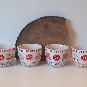Op de afbeelding: Vier witte keramieken bekers met kleurrijke illustraties van huisjes en het rode D-E logo. Elke beker heeft een unieke Nederlandse tekst langs de rand en een patroon van kleine veelkleurige stippen aan de onderkant, als een decoratieve set.