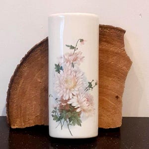 Könnte beinhalten: Eine zylindrische Keramikvase mit glatter, cremeweißer Oberfläche. Auf der Vorderseite der Vase befindet sich eine detaillierte botanische Illustration von drei hellrosa Chrysanthemenblüten mit grünen Blättern und Stielen.