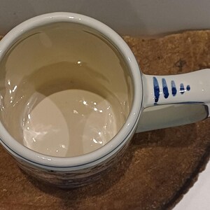 May include: A ceramic mug with a cream-colored interior and a handle featuring blue painted dash accents. The exterior displays a detailed brown and blue landscape scene with buildings and trees, finished with a smooth, glossy glaze throughout.