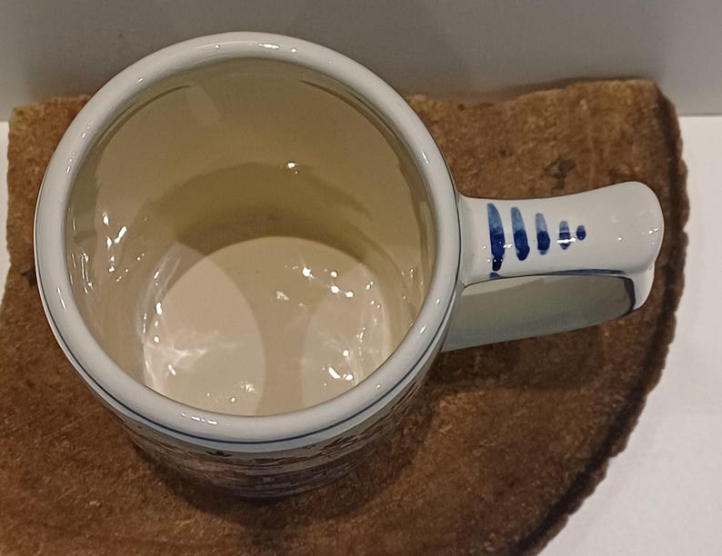 May include: A ceramic mug with a cream-colored interior and a handle featuring blue painted dash accents. The exterior displays a detailed brown and blue landscape scene with buildings and trees, finished with a smooth, glossy glaze throughout.