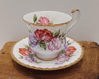 May include: A white teacup and saucer set with a floral design. The cup and saucer are trimmed with gold and feature red, pink, and purple flowers with green leaves. The set is placed on a wooden surface.