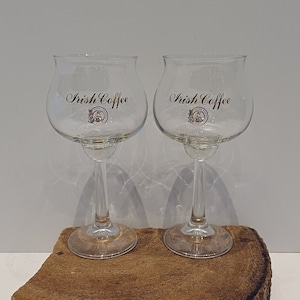 Op de afbeelding: Twee glazen Irish coffee glazen met gouden letters die "Irish Coffee" lezen. De glazen staan op een houten oppervlak.