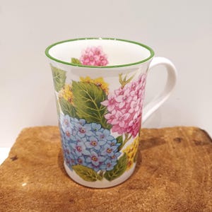 May include: A white ceramic mug with a green rim and a floral design featuring pink, blue, and yellow hydrangeas.