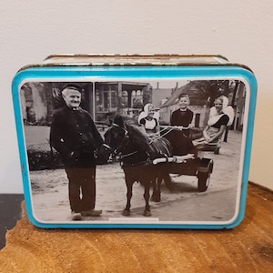 Può includere: Una scatola di latta vintage con un coperchio blu e una foto in bianco e nero di un carro trainato da un cavallo con quattro persone in abiti tradizionali olandesi. Il testo "KLEDERDRACHTEN WALCHEREN" è stampato sul fondo della scatola.