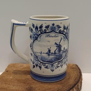 May include: A ceramic beer mug featuring a white base with blue floral patterns and a central illustration of a Dutch landscape with a windmill and the text Heineken. The mug has a sturdy handle and a blue band around the base and rim.