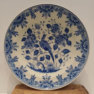 May include: A round, white ceramic plate with a blue floral and bird design. The plate features a central image of a bird perched on a branch, surrounded by blue flowers and foliage. The rim is decorated with a repeating floral pattern.