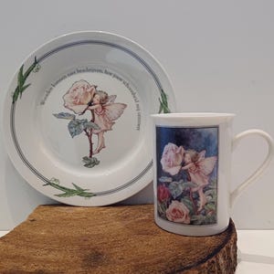 Könnte beinhalten: Ein weißes Porzellan-Teetassen- und Untertassen-Set mit einem Blumendesign, das eine Fee und Rosen zeigt. Die Untertasse trägt den Text "Words can not describe, how your beauty, my heart, converts".