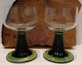 May include: Two clear wine glasses with green stems and bases. The stems have a ribbed design. The bases are a lighter shade of green. The glasses are in front of a textured, brown background.