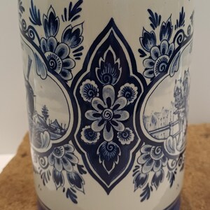 May include: A cylindrical ceramic mug featuring a cream-colored base with intricate dark blue floral patterns and circular vignettes depicting traditional Dutch landscapes, including a windmill and canal scene, with a solid blue band at the base.