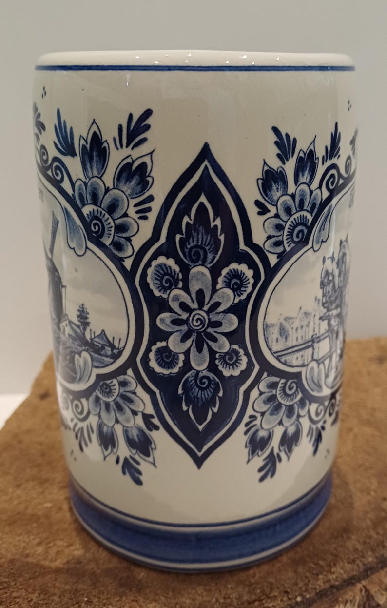 May include: A cylindrical ceramic mug featuring a cream-colored base with intricate dark blue floral patterns and circular vignettes depicting traditional Dutch landscapes, including a windmill and canal scene, with a solid blue band at the base.