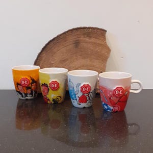 Peut inclure: Quatre tasses en céramique avec des illustrations de dessins animés colorés et les lettres "D-E" sur un emblème rouge. Les tasses ont des motifs différents, avec des personnages et des motifs, avec une anse sur une tasse. Les tasses sont sur une surface sombre.