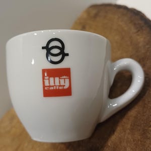 以下が含まれることがあります： 白い陶器製のエスプレッソカップ。黒い円形のロゴの下に、白い文字で「illy caffe」と書かれた赤い四角いマークがデザインされています。カップは滑らかで光沢のある仕上げで、右側にはコーヒーを持つための湾曲したハンドルが付いています。