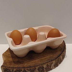 Pode incluir: Um porta-ovos de cerâmica rosa claro com quatro ovos castanhos. O porta-ovos tem um design curvo com compartimentos individuais para cada ovo. O porta-ovos é colocado sobre uma superfície de madeira.