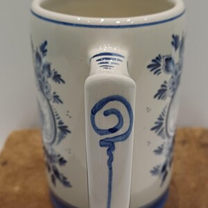 May include: A cylindrical ceramic mug with a cream-colored base and blue hand-painted floral motifs. The handle features a blue spiral design, and the rim and base are accented with thin blue horizontal lines, creating a classic decorative appearance.