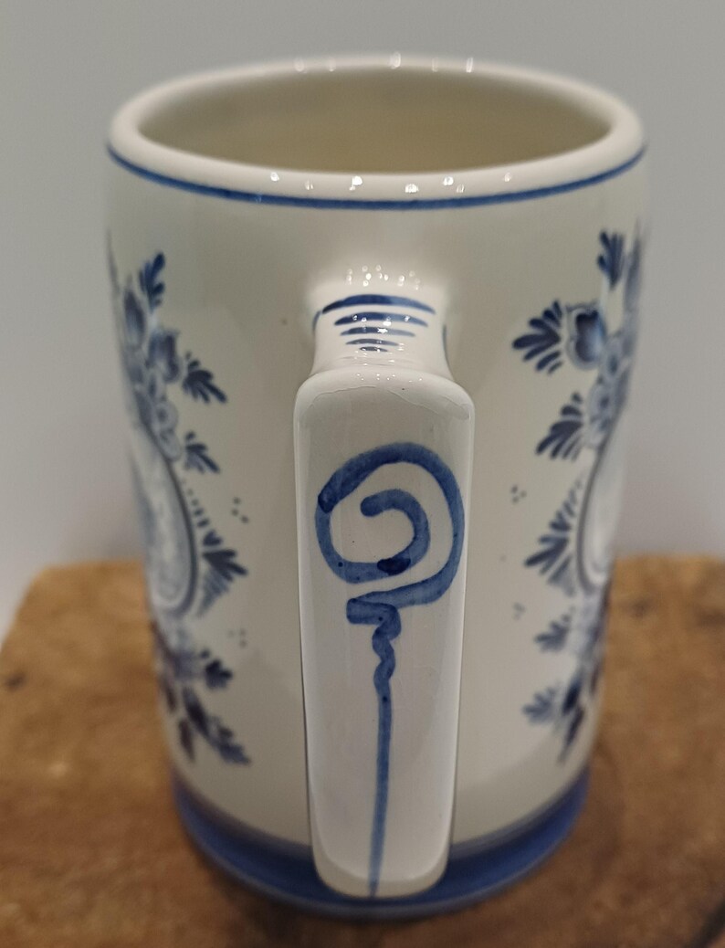 May include: A cylindrical ceramic mug with a cream-colored base and blue hand-painted floral motifs. The handle features a blue spiral design, and the rim and base are accented with thin blue horizontal lines, creating a classic decorative appearance.