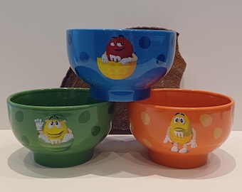 以下が含まれることがあります： M&M'sのキャラクターが描かれたセラミック製ボウル3個セット。上部の青いボウルには赤いキャラクター、左下の緑のボウルには黄色いキャラクター、右下のオレンジのボウルには水玉模様の黄色いキャラクターが描かれています。