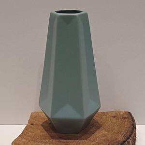 Op de afbeelding: Een matte saliegroene keramische vaas met een geometrisch, veelzijdig ontwerp. De vaas heeft een taps toelopend, zeshoekig lichaam met scherpe, hoekige randen en een brede basis die smaller wordt naar de ronde opening aan de bovenzijde.