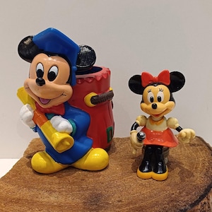 May include: A red, yellow, and blue plastic Mickey Mouse figurine wearing a graduation cap and holding a yellow and red scroll. A smaller Minnie Mouse figurine wearing a red dress and holding a microphone stands next to Mickey Mouse.