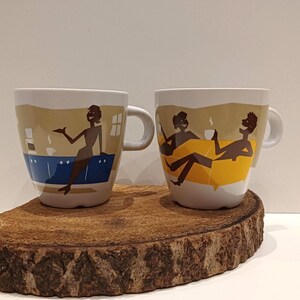 Peut inclure: Deux tasses en céramique blanche ornées de silhouettes brunes stylisées. La tasse de gauche montre une personne debout près d'un comptoir bleu. La tasse de droite représente deux personnes sur un canapé jaune, l'une tenant une tasse.