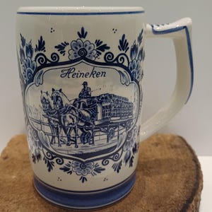 May include: A cream-colored ceramic beer mug featuring a blue transferware-style design. The central graphic depicts a horse-drawn carriage with a driver and the text Heineken. The mug is decorated with ornate blue floral patterns and borders.