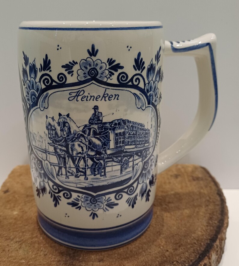 May include: A cream-colored ceramic beer mug featuring a blue transferware-style design. The central graphic depicts a horse-drawn carriage with a driver and the text Heineken. The mug is decorated with ornate blue floral patterns and borders.
