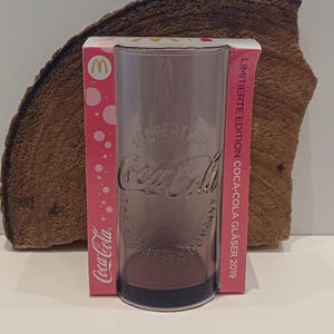 May include: A clear Coca-Cola glass with the Coca-Cola logo and text. The glass is in its original pink packaging, which has the McDonald's logo. The packaging reads "LIMITIERTE EDITION COCA-COLA GLÄSER 2019".