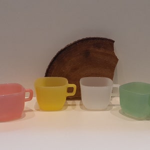 May include: Four pastel-coloured, square-shaped glass cups: pink, yellow, white, and green. Each cup has a small handle. A piece of wood is behind the cups.