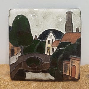 May include: Square ceramic tile with a landscape scene. The artwork shows a bridge, buildings, trees, and a tower, in brown, green, and white. The tile has a glossy finish and an irregular shape. The scene evokes a quaint village.
