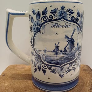 May include: A white ceramic beer mug featuring blue floral patterns and a central illustration of Dutch windmills near a body of water. The word Heineken is printed in blue script above the landscape scene. The mug has a sturdy handle and a blue base.