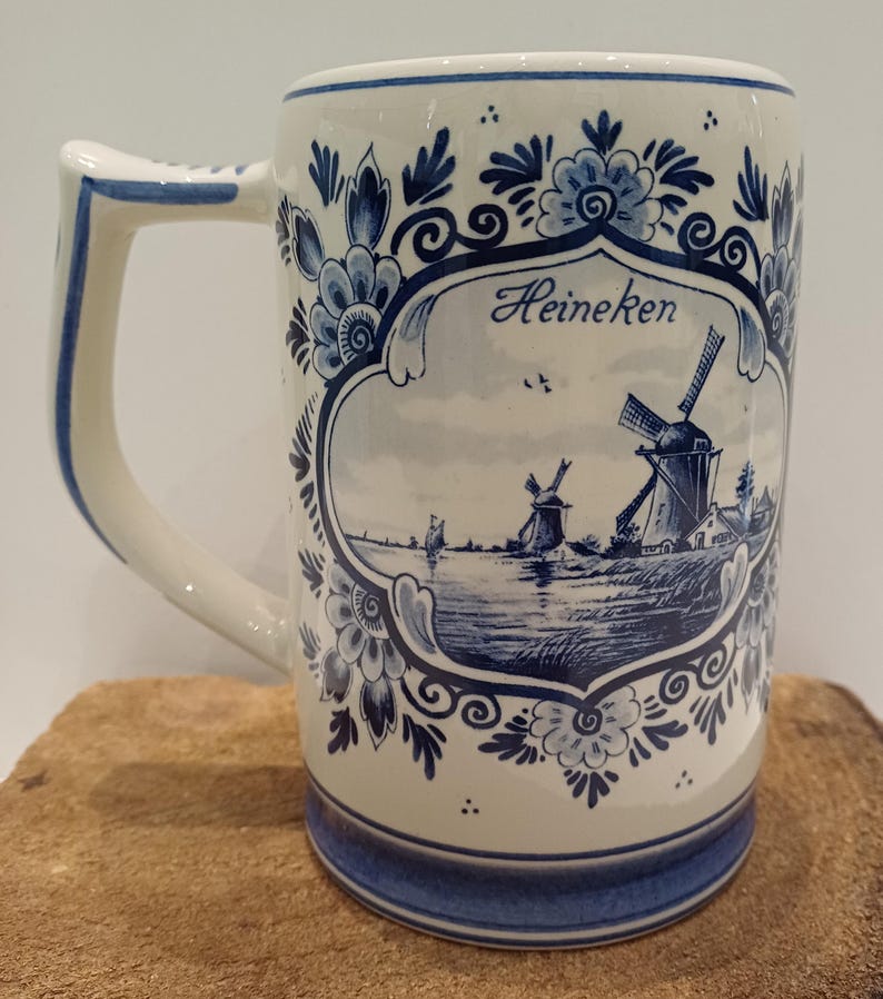 May include: A white ceramic beer mug featuring blue floral patterns and a central illustration of Dutch windmills near a body of water. The word Heineken is printed in blue script above the landscape scene. The mug has a sturdy handle and a blue base.